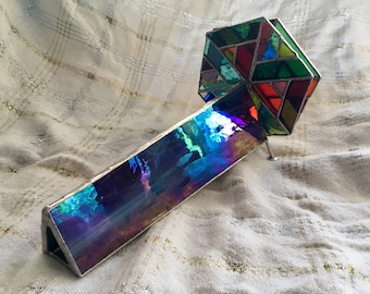 May include: A kaleidoscope constructed from iridescent glass panels and a multi-colored stained glass end piece. The body features a reflective, color-shifting surface, while the hexagonal end is composed of various geometric glass shards.