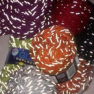Op de afbeelding: Assortiment van garenklossen in verschillende kleuren, waaronder paars, oranje, groen en grijs. Het garen heeft een reflecterende draad erin geweven, waardoor een glinsterend effect ontstaat. Het etiket luidt "Glow Reflect with 27% Superwash".