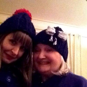 Op de afbeelding: Twee vrouwen dragen blauwe gebreide mutsen. De ene muts heeft een rode pompon en de andere een witte en grijze strik. De vrouwen lachen en kijken naar de camera.