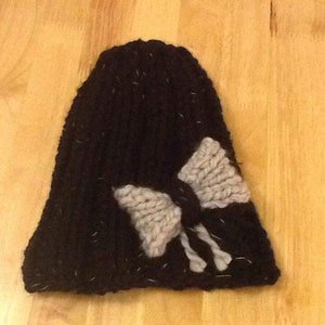 Op de afbeelding: Zwarte gebreide beanie met een grote grijze strik aan de voorkant.