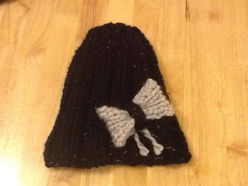 Op de afbeelding: Zwarte gebreide beanie met een grote grijze strik aan de voorkant.