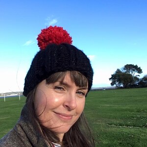 Op de afbeelding: Een vrouw draagt een zwarte gebreide beanie met een rode pompon. Ze staat in een veld met groen gras en een blauwe lucht op de achtergrond.