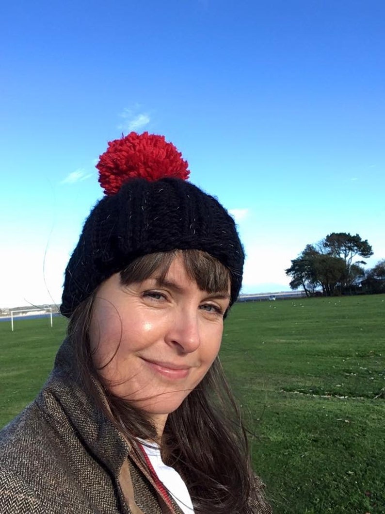 Op de afbeelding: Een vrouw draagt een zwarte gebreide beanie met een rode pompon. Ze staat in een veld met groen gras en een blauwe lucht op de achtergrond.