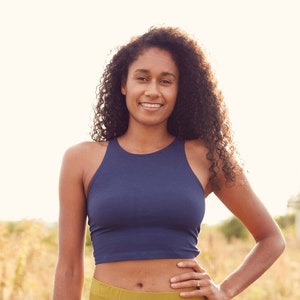 May include: A woman wearing a navy blue crop top and mustard yellow leggings. She is standing in a field of tall grass.