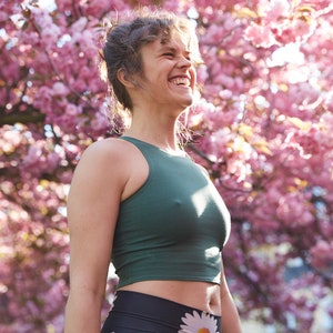 May include: A woman wearing a green tank top and black leggings with a floral print stands in front of a pink cherry blossom tree. She is smiling and looking up.