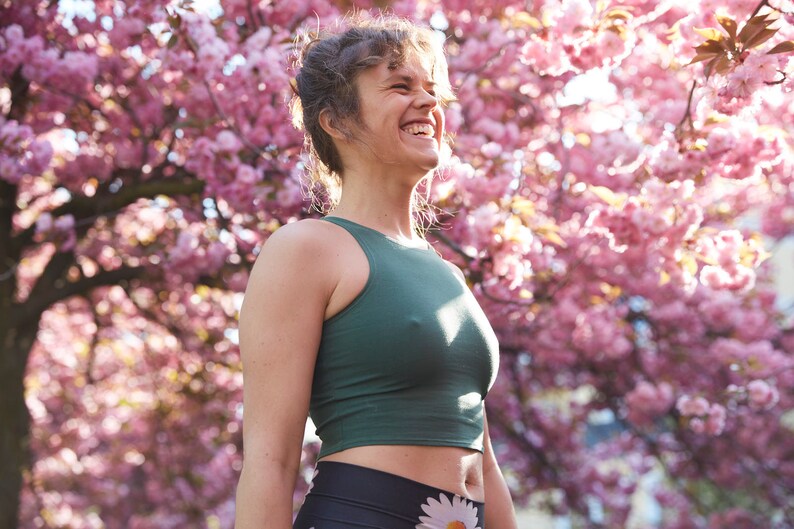 May include: A woman wearing a green tank top and black leggings with a floral print stands in front of a pink cherry blossom tree. She is smiling and looking up.