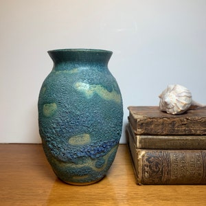 May include: A blue and green ceramic vase with a textured surface. The vase has a rounded shape and a wide opening. It is sitting on a wooden surface.