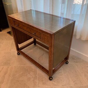 May include: A rectangular wooden desk featuring a smooth, light-colored top surface and a single drawer with a small metal knob. The desk is supported by a sturdy frame with four small wheels at the base, allowing for mobility across the floor.