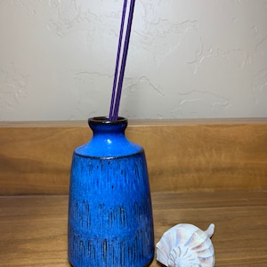 May include: A blue ceramic vase with a dark rim holds three purple incense sticks. A white and tan seashell rests beside the vase on a wooden surface. The vase has a textured, glazed finish.