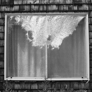 May include: A black and white photograph of a window with a broken pane. The window is in a wooden frame and is set into a wooden wall. The broken pane is covered with a torn piece of material.