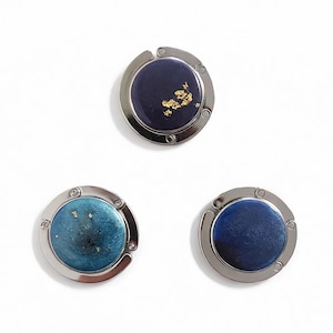 May include: Three silver-coloured purse hangers with circular tops. The tops feature dark blue, teal, and navy blue resin with gold flecks. The hangers are designed to hold purses and bags.