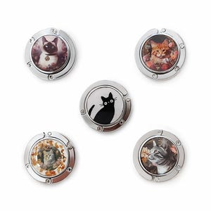 Op de afbeelding: Een verzameling van vijf zilverkleurige tashangers, elk met een ander kattenportret. De kattenafbeeldingen zijn geplaatst tegen bloemrijke achtergronden, waarvan één een zwart katten silhouet toont. Deze accessoires zijn ontworpen om handtassen op te hangen.