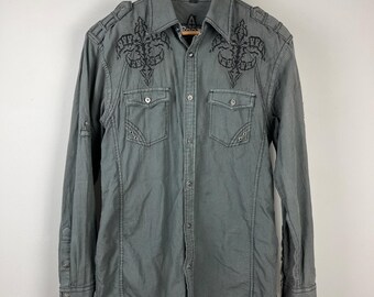 May include: Gray button-down shirt with a small checkered pattern. The shirt features black embroidered designs on the chest and shoulders, two front pockets, and long sleeves. The shirt is hanging on a wooden hanger.