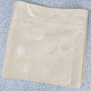 May include: Cream-colored fabric featuring a repeating pattern of smiling suns, crescent moons, and stars. The celestial design is woven into the fabric, creating a subtle texture. The fabric is folded, showing the pattern's repeat.