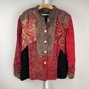 May include: A patchwork jacket with a high collar and long sleeves. The jacket features a mix of red, black, and gold patterned fabrics. It has gold buttons down the front and is hanging on a wooden hanger.