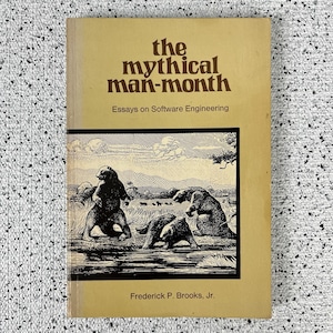May include: A paperback book titled The Mythical Man-Month: Essays on Software Engineering by Frederick P. Brooks, Jr. The cover is tan with a black and white illustration of three bears in a landscape near a body of water at the bottom.