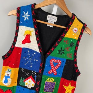 May include: A colorful, festive holiday vest with a patchwork design. The vest features various Christmas-themed appliqués, including a snowman, snowflakes, and a candy cane. The vest is made of a knitted material and has a black back and red stitching.