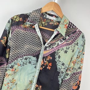 May include: A long-sleeved button-down shirt featuring a multi-colored patchwork pattern in shades of black, sage green, and orange. The garment displays a mix of floral motifs, geometric grids, and paisley-like designs with a collar and snap buttons.