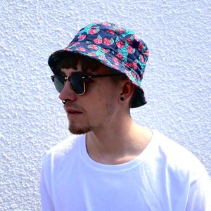 Könnte beinhalten: Eine Person trägt einen blauen und pinkfarbenen Bucket-Hut mit einem Wassermelonenmuster. Der Hut hat eine breite Krempe und ist aus einem leichten Stoff gefertigt.