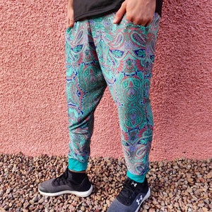 Puede incluir: Pantalones deportivos con estampado de cachemira en verde azulado, rojo y morado. Los pantalones deportivos tienen puños turquesa en los tobillos y bolsillos. La persona lleva zapatillas deportivas negras y grises. El fondo es una pared rosa texturizada y pequeñas piedras.