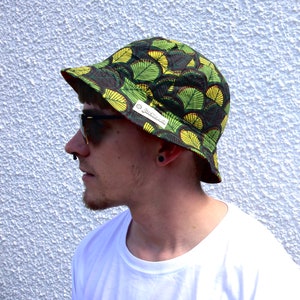 Könnte beinhalten: Eine Person trägt einen grünen und gelben Bucket Hat mit einem Blatt-Muster. Der Hut hat ein weißes Etikett mit dem Text "Jackskull" darauf. Die Person trägt auch eine Sonnenbrille.