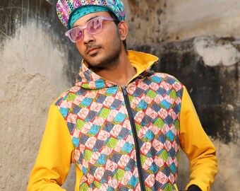 May include: A yellow and black hooded sweatshirt with a colourful geometric pattern on the front. The hood is up and the zipper is closed. The person is wearing a colourful patterned hat and sunglasses.