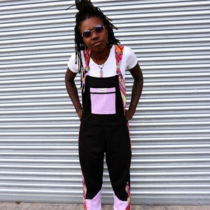 May include: Black overalls with a light purple pocket and cuffs, accented with colorful patterned fabric on the straps and legs. The overalls are paired with a white t-shirt and black shoes. The overall design is a unique and stylish fashion statement.