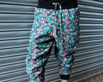 May include: Colorful patterned pants with a black waistband and cuffs. The pants feature a teal background with a repeating pattern of pink and green shapes. The drawstring is black. The pants are cropped at the calf.