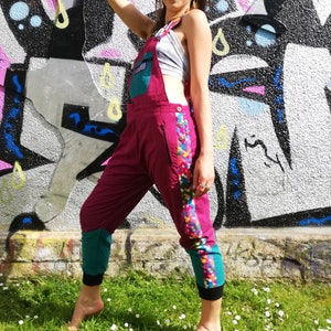 Puede incluir: Una mujer lleva un mono rosa con una raya colorida a lo largo del lateral. El mono tiene un ribete turquesa y una cintura negra. Está de pie frente a una pared con grafitis y lleva gafas de sol.