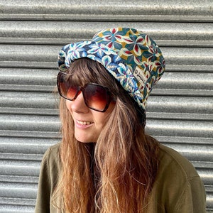 Op de afbeelding: Een vrouw draagt een groene shirt en een zonnebril met een bloemenprint bucket hat. De hoed heeft een klein label met de tekst "Duck and Cover".