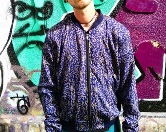 May include: A blue bomber jacket with a gold floral pattern, black zipper, and navy blue cuffs. The person in the image is wearing a patterned hat and sunglasses. The background features colorful graffiti art.