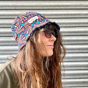 Könnte beinhalten: Eine Frau trägt einen schwarzen Bucket-Hut mit einem bunten Blumenmuster und einem goldenen Etikett mit der Aufschrift "Jackalope". Sie trägt auch eine Sonnenbrille.