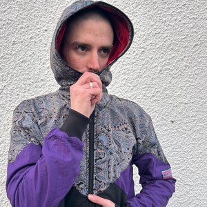 Könnte beinhalten: Eine schwarze, violette und silberne Kapuzenjacke mit Paisley-Muster. Die Jacke hat einen Reißverschluss und einen kleinen Logo-Patch am Ärmel.