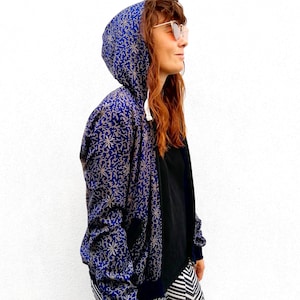 May include: A blue and gold patterned hooded jacket with a zip closure. The jacket is being worn by a person with long brown hair and sunglasses.