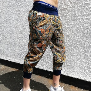 Puede incluir: Pantalones cortos con estampado de cachemira con puños y cintura azul marino. Los pantalones presentan un patrón colorido con tonos de naranja, verde azulado y marrón. La persona lleva zapatillas blancas con detalles coloridos.