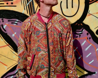 May include: A red, gold, and blue paisley patterned bomber jacket with a zipper closure. The jacket has a red ribbed collar and cuffs. The person is wearing a visor and sunglasses.