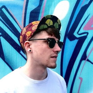 May include: A person wearing a colourful patterned baseball cap with a green, yellow, red and orange design. The person is also wearing sunglasses and a white t-shirt.