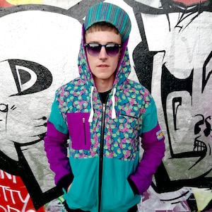 May include: A turquoise, purple, and white patterned zip-up hoodie with a black hood. The hoodie has a white zipper and a purple pocket on the left side. The model is wearing a striped hat and sunglasses.