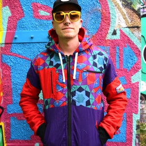 May include: A person wearing a colorful geometric patterned jacket with a black hat and yellow sunglasses. The jacket has a zipper and a hood. The person is standing in front of a graffiti wall.