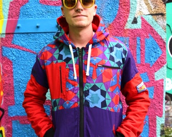 May include: A person wearing a colourful geometric patterned jacket with a black hat and yellow sunglasses. The jacket has a zipper and a hood. The person is standing in front of a graffiti wall.