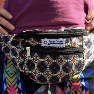 May include: A patterned fanny pack with a geometric design in red, gray, and blue. The bag has two zippered compartments and a black strap. The brand name "jackFruit" is displayed on a white label.