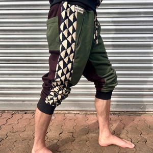 May include: Green, burgundy, and black sweatpants with a geometric design. The trousers feature a black and white diamond pattern down the side, a zipped pocket, and black cuffs. The person is standing barefoot in front of a corrugated metal wall.