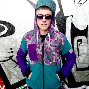 May include: A teal, purple, and pink patterned hooded jacket with a zipper closure. The jacket has a purple pocket on the left side and a white drawstring hood. The model is wearing a striped hat and sunglasses.