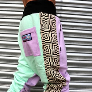 May include: A pair of multi-colored sweatpants with a black waistband and a zipper on the side. The sweatpants have a light green, purple, and black and white geometric pattern. The purple section has a pocket with a logo that says "SPORT" with a flower design.