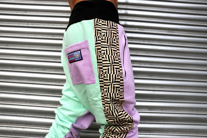 May include: A pair of multi-colored sweatpants with a black waistband and a zipper on the side. The sweatpants have a light green, purple, and black and white geometric pattern. The purple section has a pocket with a logo that says "SPORT" with a flower design.