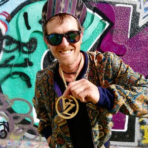 May include: A man wearing a patterned cap, sunglasses, and a paisley patterned jacket is holding a gold pendant with the letters "VZ" on it. The pendant is on a gold chain.