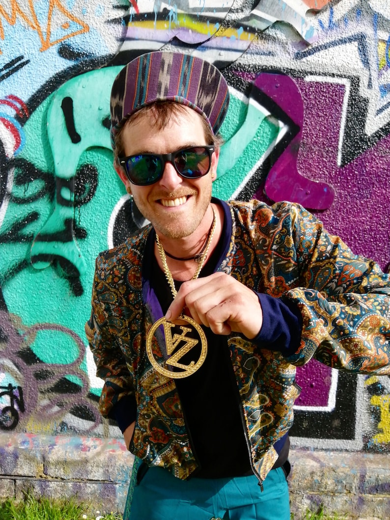 May include: A man wearing a patterned cap, sunglasses, and a paisley patterned jacket is holding a gold pendant with the letters "VZ" on it. The pendant is on a gold chain.
