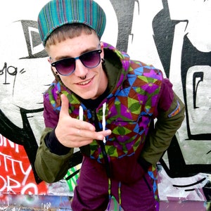 May include: A person wearing a green and purple geometric patterned jumpsuit with a blue and green striped hat and sunglasses. The person is standing in front of a graffiti wall.