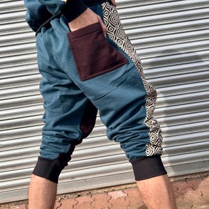 May include: Teal blue cropped pants with a brown patch pocket and a geometric pattern on the side. The pants have a black elastic waistband and cuffs.
