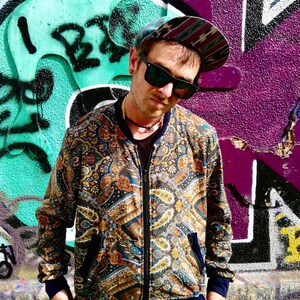May include: A man wearing a paisley patterned bomber jacket with a zipper closure. The jacket has a blue, gold, and brown color scheme. He is wearing a blue and black patterned hat and sunglasses.